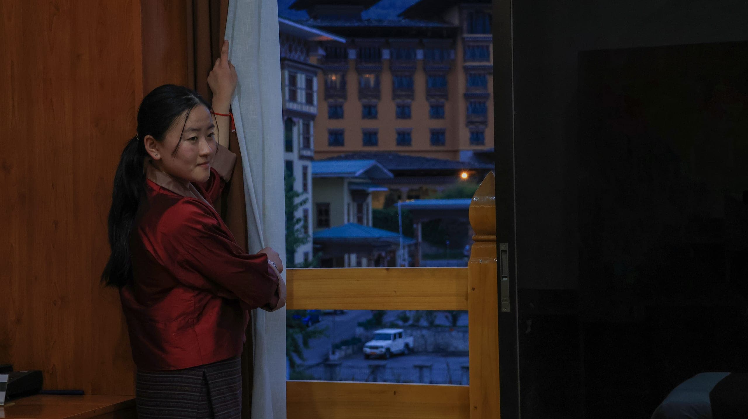 A staff member in traditional dress opening the curtains to an evening view of Thimphu